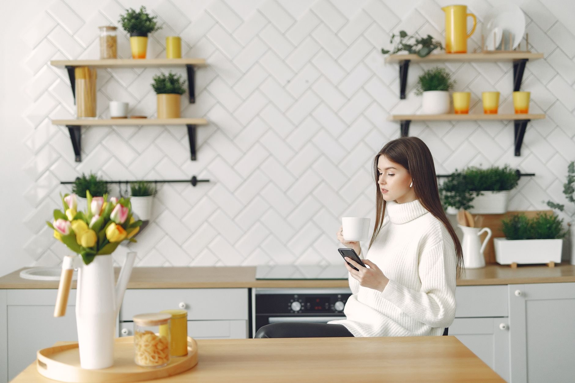 Side view of focused young woman in warm sweater browsing mobile phone in cozy kitchen